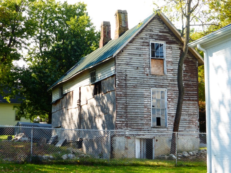 The back and left sides of the house (facing south and east, respectively).  Note the fact that the back of the house is covered with thin sheets of wood and the original siding is gone.
